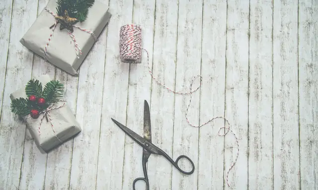 Twee in grijs papier ingepakte cadeaus met rood-wit touw en dennentakjes, naast een schaar en een klosje touw op een lichte houten vloer