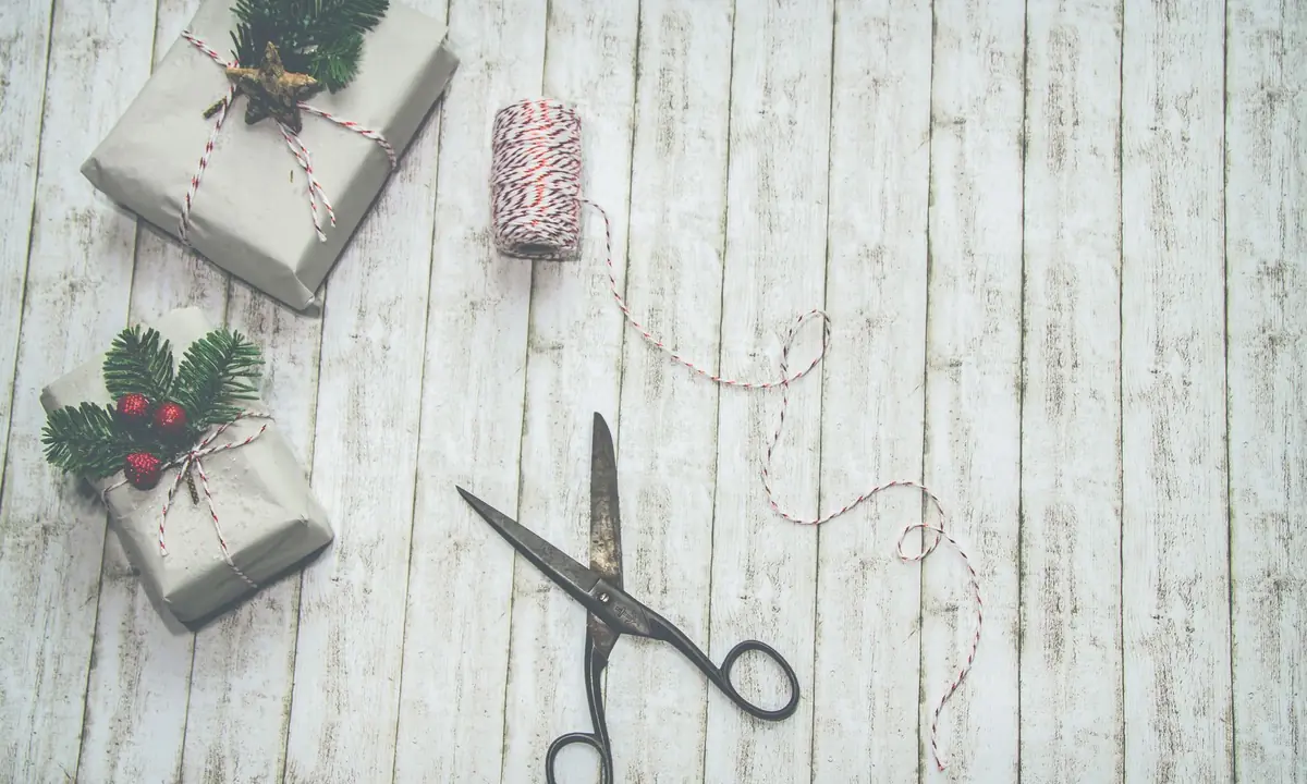 Twee in grijs papier ingepakte cadeaus met rood-wit touw en dennentakjes, naast een schaar en een klosje touw op een lichte houten vloer