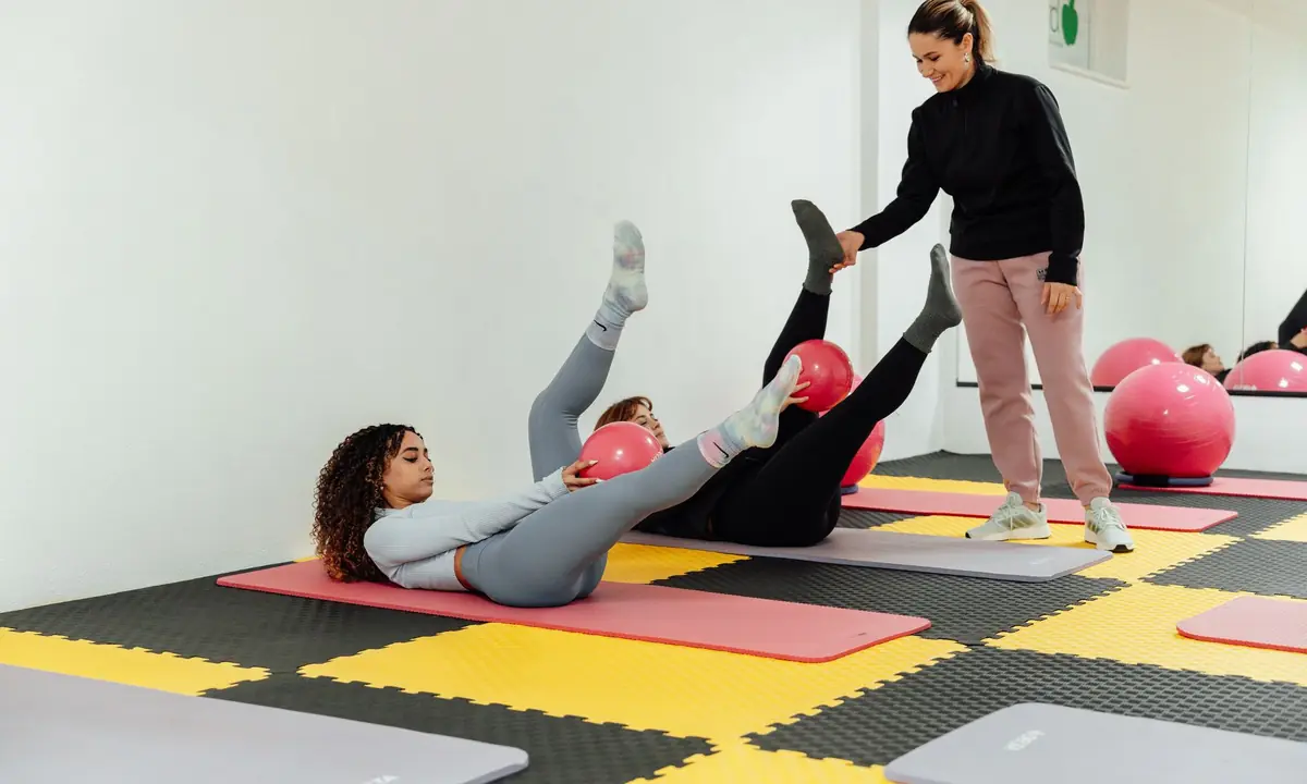Twee vrouwen liggen op hun rug op een mat met een been omhoog, terwijl een instructeur een been ondersteunt tijdens een matles pilates